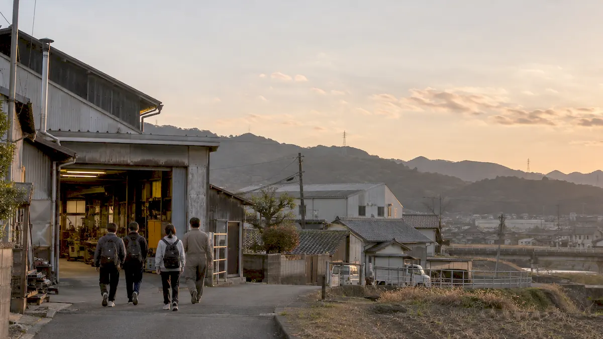 夕方の町で、若者たちが小さな工場へ向かって歩いている後ろ姿（イメージ）