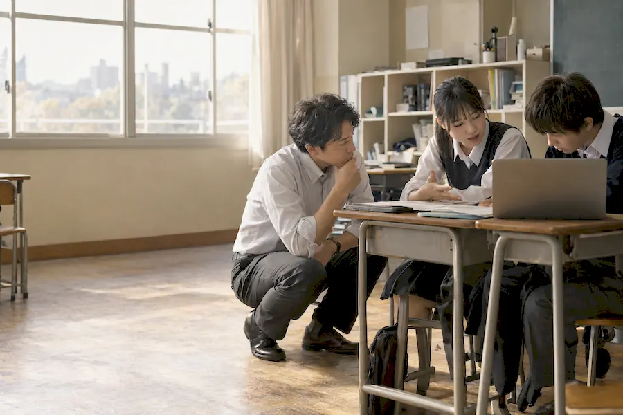 教室で、先生が机のそばにしゃがみ、生徒二人の話を聞いている（イメージ）